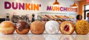 A close-up shot of six different Dunkin' Munchkins donut holes—glazed, chocolate glazed, cinnamon sugar, powdered sugar, jelly-filled, and butternut,lined up on a reflective counter with a bright Dunkin' shop interior in the background.