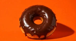 A person holding a fresh Dunkin glazed chocolate donut in a branded paper bag inside a modern Dunkin store with coffee cups, donut displays, and workers in the background.